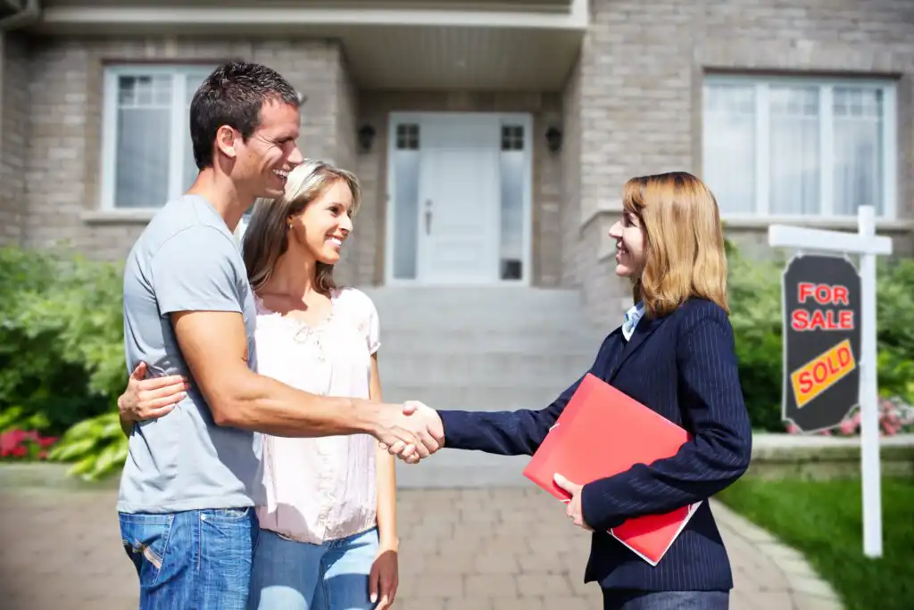 A smiling couple stands in front of a house, shaking hands with a real estate agent holding a red folder. A For Sale sign with a Sold sticker is visible in the yard.
