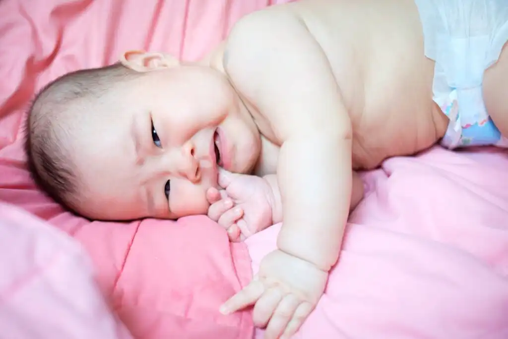 A baby wearing a diaper lies on a pink blanket, appearing to cry or be upset, with one hand near their mouth and the other hand resting on the blanket.