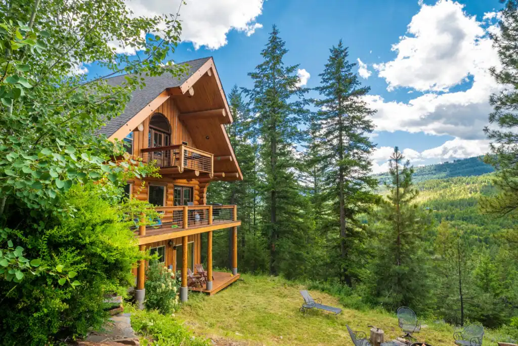 A wooden cabin with balconies is surrounded by tall pine trees and lush greenery, overlooking a scenic forested valley under a blue sky with scattered clouds. Outdoor chairs and a fire pit are visible on the grassy area nearby.