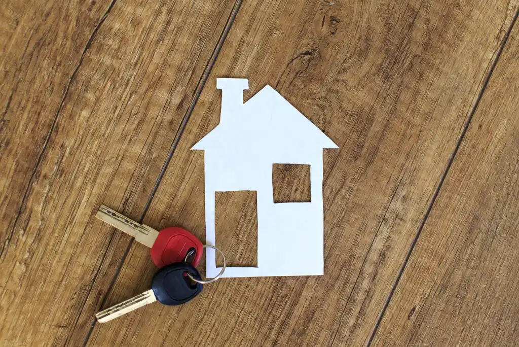 A paper cutout of a house and two keys with red and black key covers rest on a wooden surface, symbolizing home ownership or real estate.