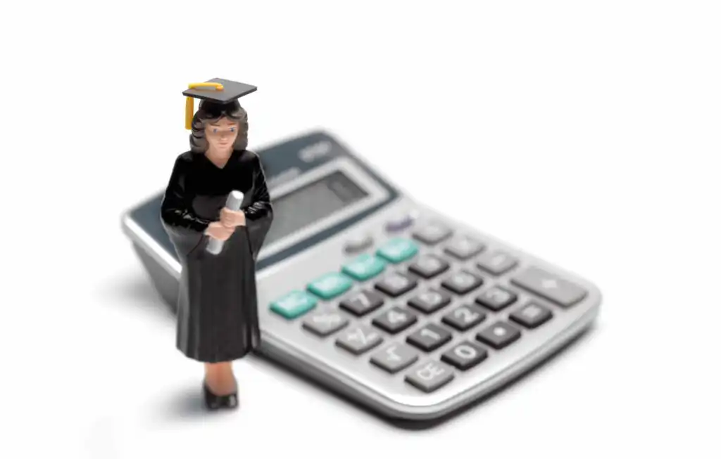 A small figurine of a graduate in a cap and gown holding a diploma stands in front of a calculator on a white background.