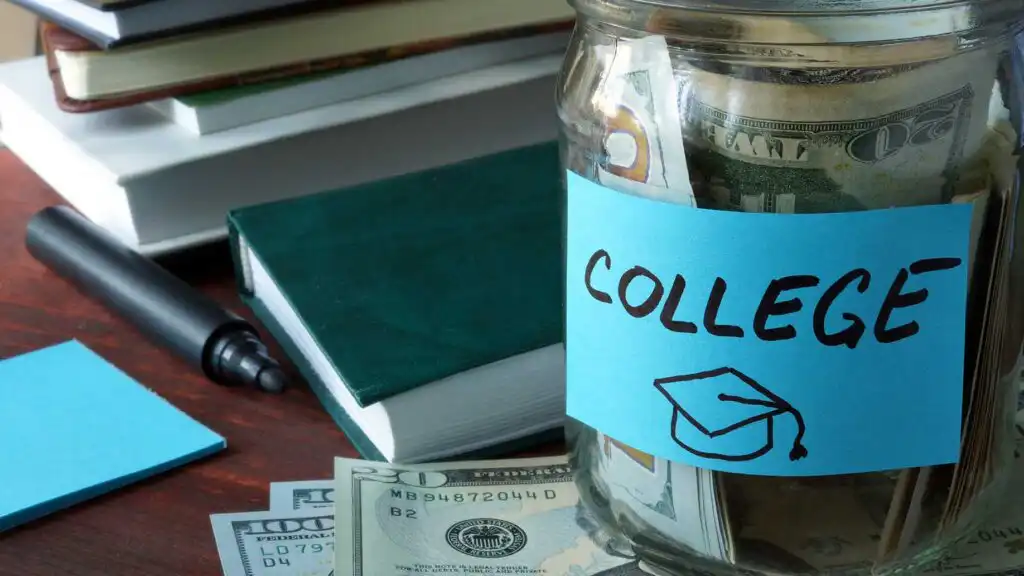 A glass jar labeled COLLEGE with a graduation cap drawing is filled with cash. The jar is on a table with books, a marker, sticky notes, and U.S. dollar bills surrounding it.