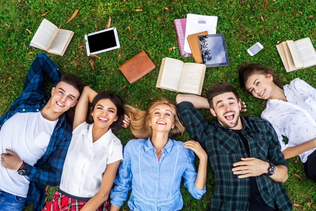 Five young adults lie on grass in a row, smiling and laughing. Books, notebooks, a tablet, and a phone are scattered nearby. They appear relaxed and happy, enjoying a break outdoors.