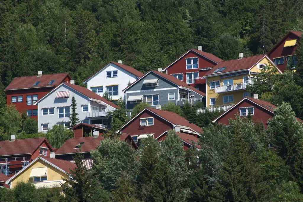Colorful wooden houses with red, yellow, white, and blue facades are nestled on a lush, green hillside surrounded by dense trees and greenery.