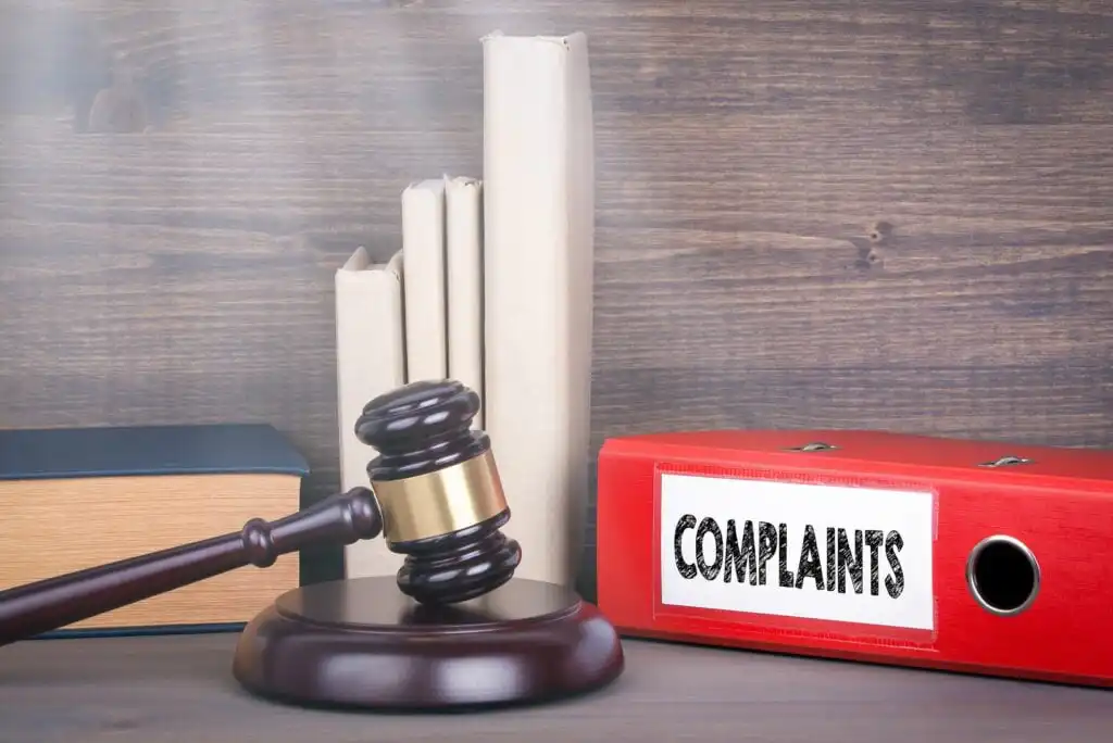 A judges gavel rests on a sound block in front of books and a red binder labeled COMPLAINTS on a wooden surface with a wood-paneled background.