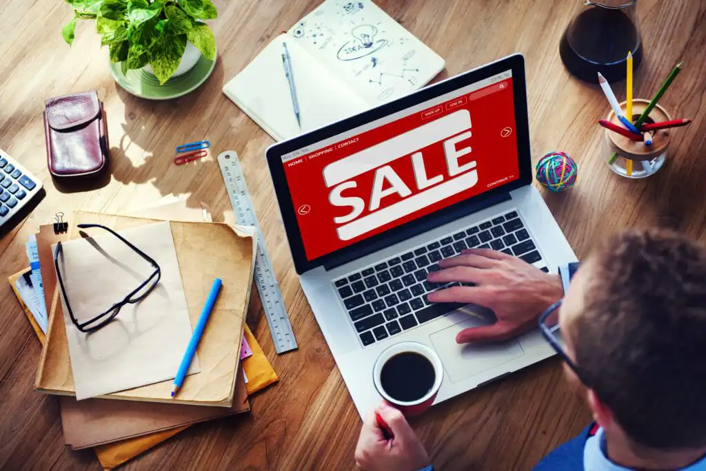 A person at a wooden desk uses a laptop displaying a large red SALE sign. The desk has a notebook, envelopes, smartphone, coffee cup, ruler, potted plant, glasses, and various office supplies.