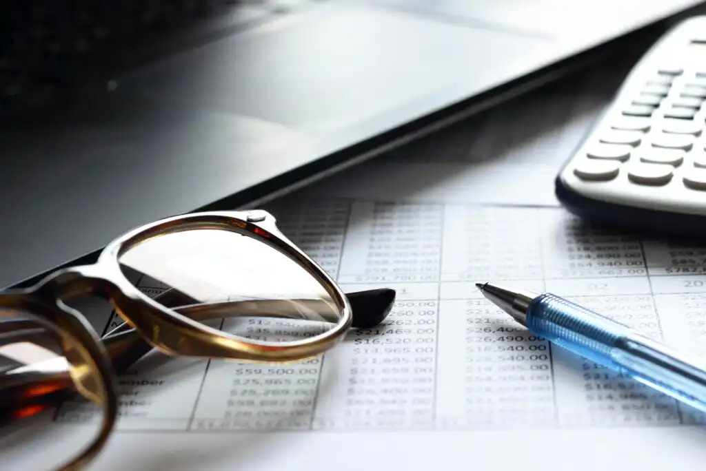 Close-up of eyeglasses, a pen, and a calculator on top of a financial spreadsheet, with part of a laptop keyboard visible in the background.