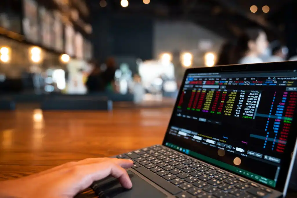 A person uses a laptop displaying a colorful stock market trading screen while sitting at a wooden table in a modern, softly lit café. The background is blurred with warm lighting and people.