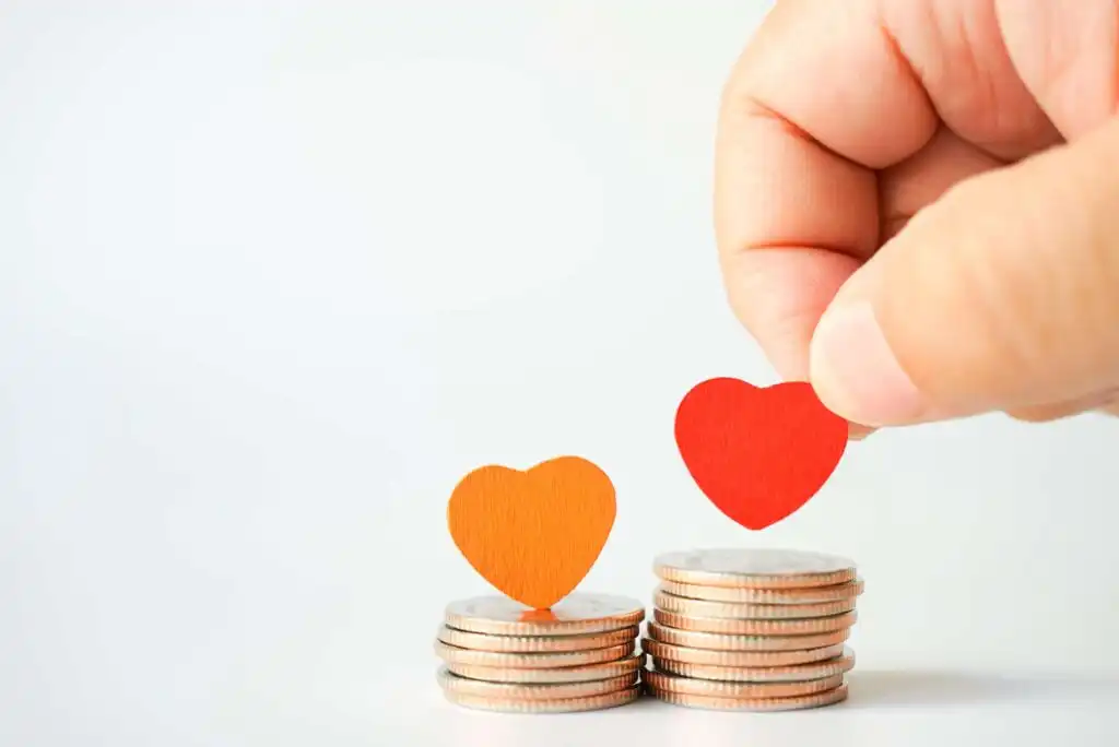 A hand places a red heart shape on a stack of coins, next to another stack topped with an orange heart, symbolizing charity and financial donations.