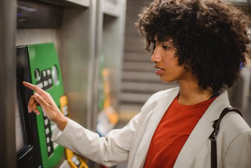 A woman with curly hair uses a ticket machine, pressing a button on the screen. She wears a light blazer over a red shirt and carries a bag over her shoulder.
