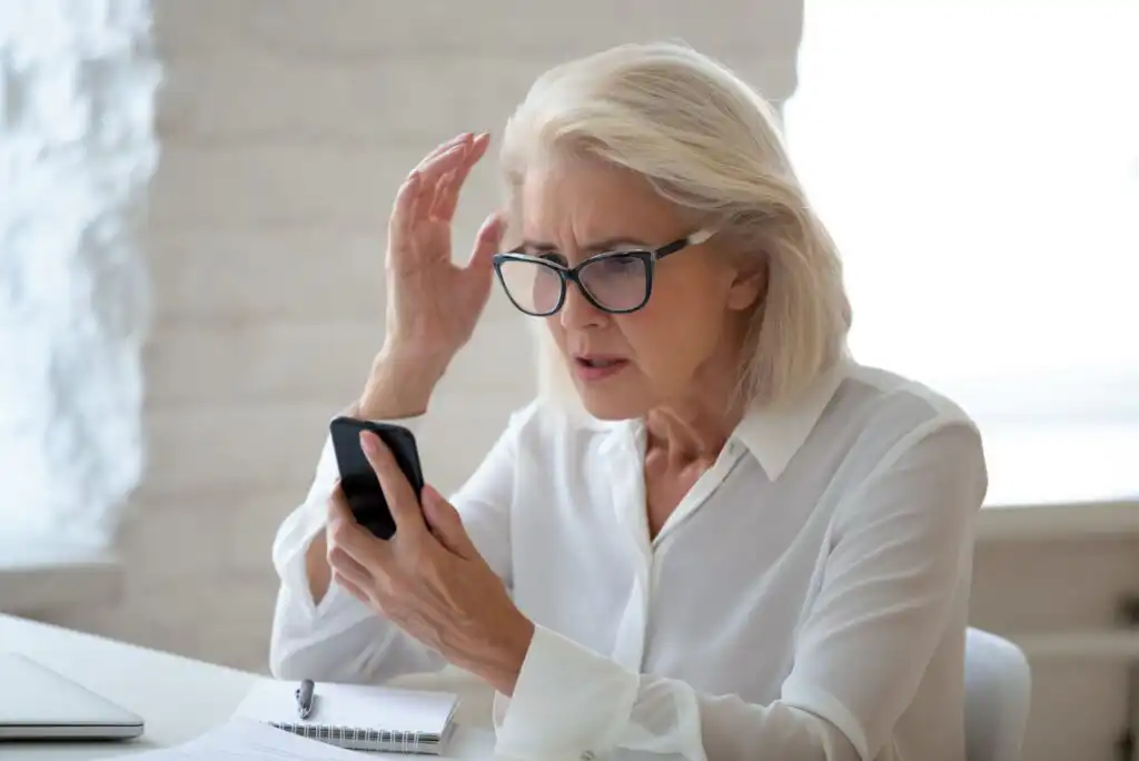 An older woman with gray hair and glasses looks confused or concerned while staring at her smartphone. She is sitting at a desk with a notebook, pen, and laptop nearby.