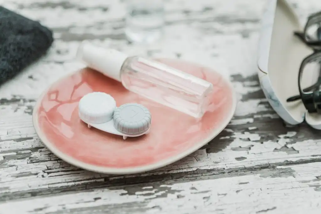 A pink dish holds a clear spray bottle and a white and blue contact lens case on a distressed white wooden surface. A pair of eyeglasses and a dark cloth are partially visible nearby.