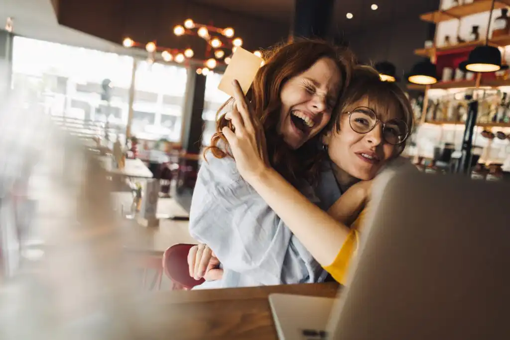 Two friends smiling and hugging while sitting at a cafe table, looking at a laptop. The atmosphere is warm and cheerful with soft lighting in the background.