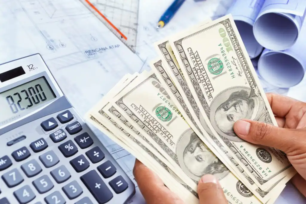 A person counts US $100 bills next to a calculator displaying 823000 and architectural blueprints, suggesting calculation of construction or project costs.