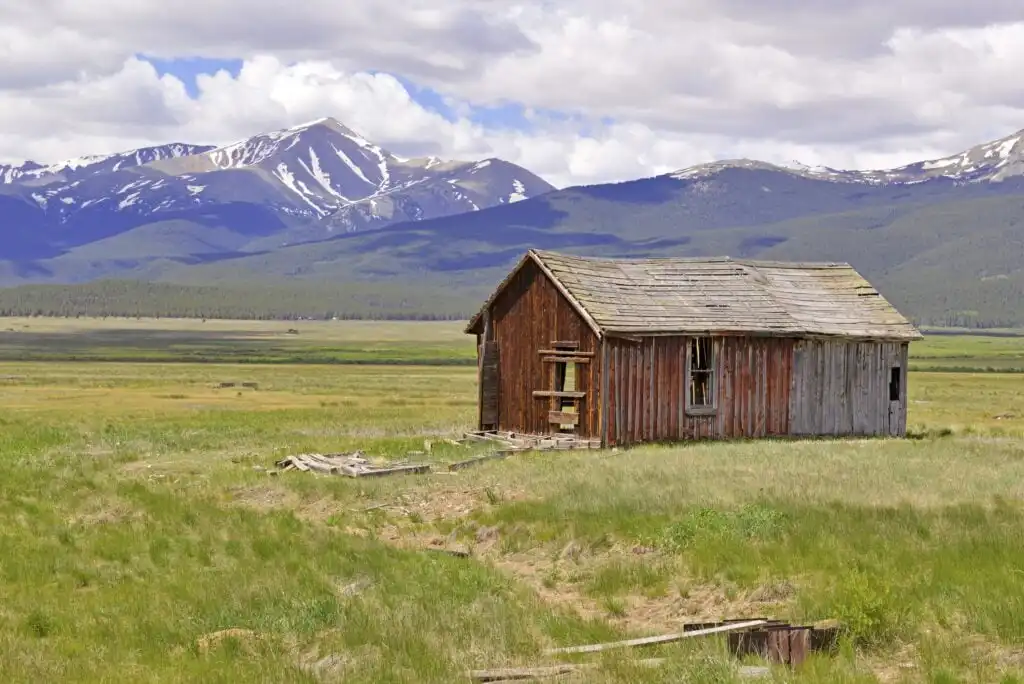 A weathered wooden cabin stands alone in a grassy field, with snow-capped mountains and a forested ridge in the background under a partly cloudy sky.