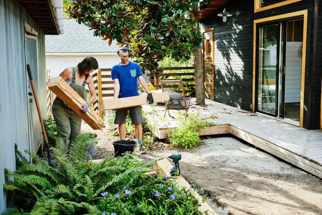 Two people work together in a backyard garden, lifting large wooden beams. They are near a house with black siding and large windows, surrounded by lush plants and a partially constructed deck.