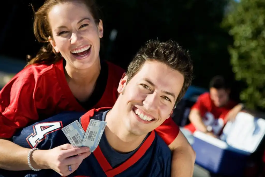 A smiling woman and man wearing sports jerseys pose outdoors. The woman holds two event tickets while hugging the man from behind. Another person in a red jersey appears blurred in the background near a cooler.