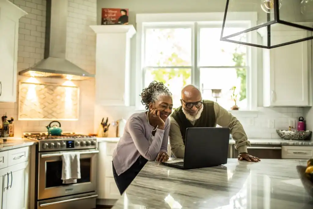 An older couple smiling and looking at a laptop together on a kitchen island in a bright, modern kitchen with white cabinets and natural light from large windows.