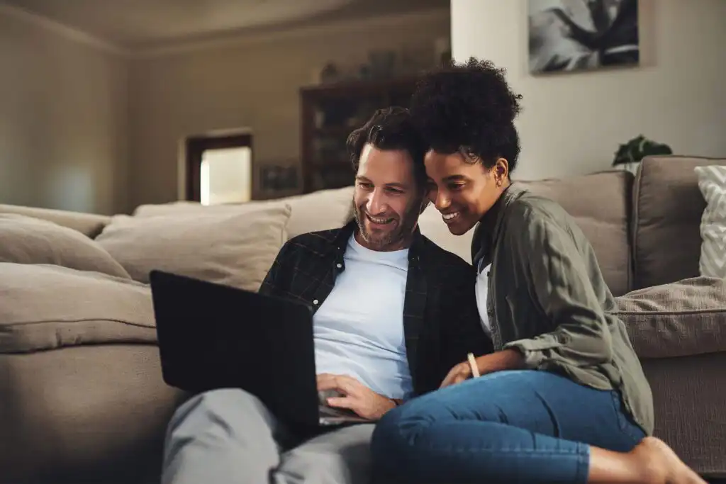 A smiling couple sits on the floor, leaning against a beige couch while looking at a laptop together in a cozy living room.