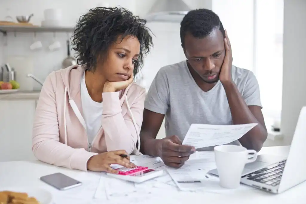 A couple sits at a table looking worried while reviewing bills and paperwork, with a laptop, coffee cup, and calculator in front of them in a bright kitchen.