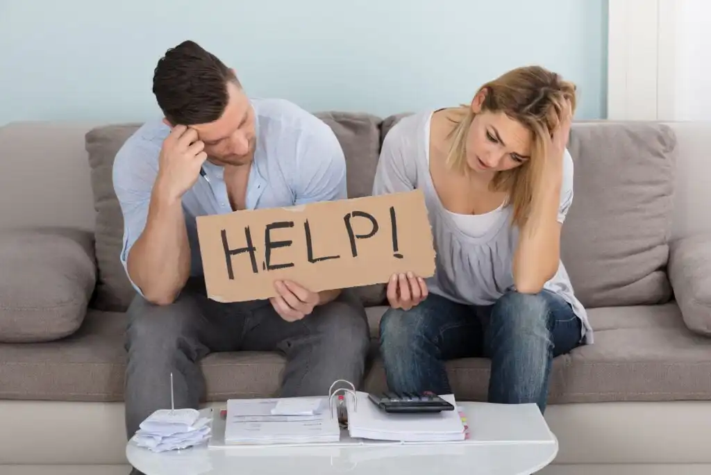A worried couple sits on a couch, holding a cardboard sign that says “HELP!” with paperwork, bills, and a calculator on the table in front of them, suggesting financial stress.