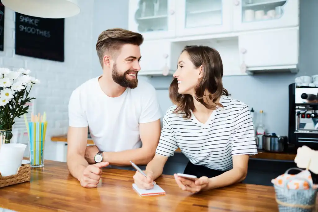 A smiling man and woman sit at a kitchen counter. The woman writes in a notebook and holds a phone, while the man leans on the counter. They are talking and appear happy in a bright, modern kitchen.