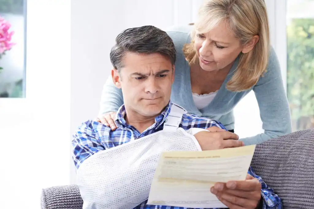 A man with an arm in a sling sits on a couch, looking concerned while reading a document. A woman stands beside him with a supportive hand on his shoulder, also looking at the paper.