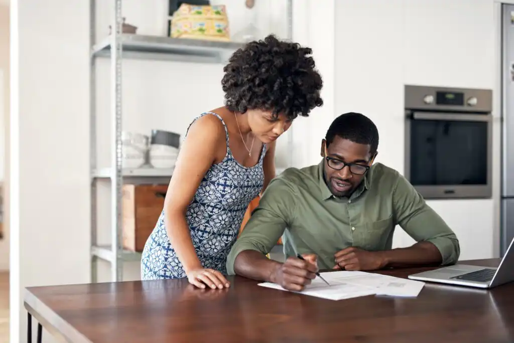 A woman and man review documents together at a table in a modern kitchen. The man writes on the papers while the woman looks on, and a laptop is open beside them.