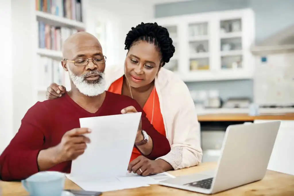 An older couple sits at a kitchen table reviewing documents together. The man holds papers while the woman stands beside him, looking at the documents. A laptop is open on the table next to them.