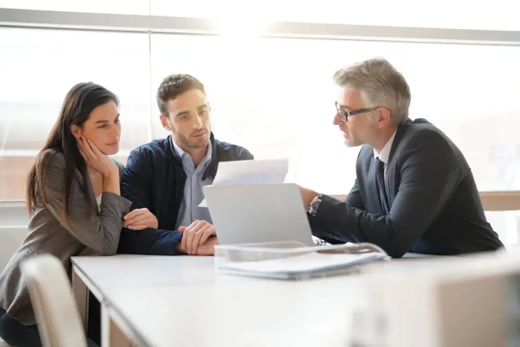 A professional man in a suit explains documents to a young couple seated across from him at a desk, with a laptop and paperwork on the table in a bright office setting.