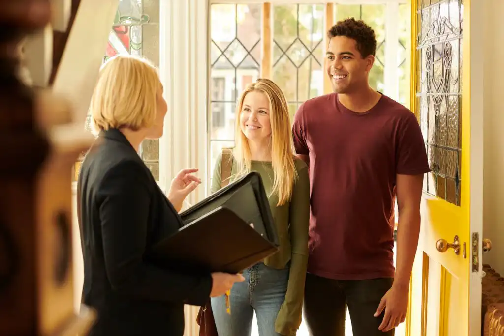 A woman holding a folder stands by the door, talking to a smiling young couple who are entering a house through a bright yellow front door. Sunlight comes through the windows behind them.