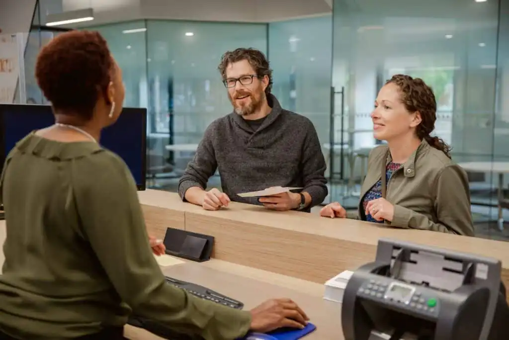 A man and a woman stand at a reception desk, smiling and holding documents, while a receptionist in a green blouse speaks to them in a modern office setting.