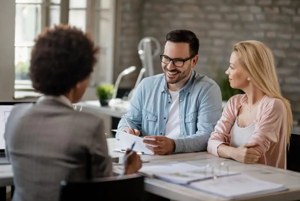 A man and woman sit together at a desk, smiling and talking with a professional across from them in an office setting, suggesting a business meeting or consultation.