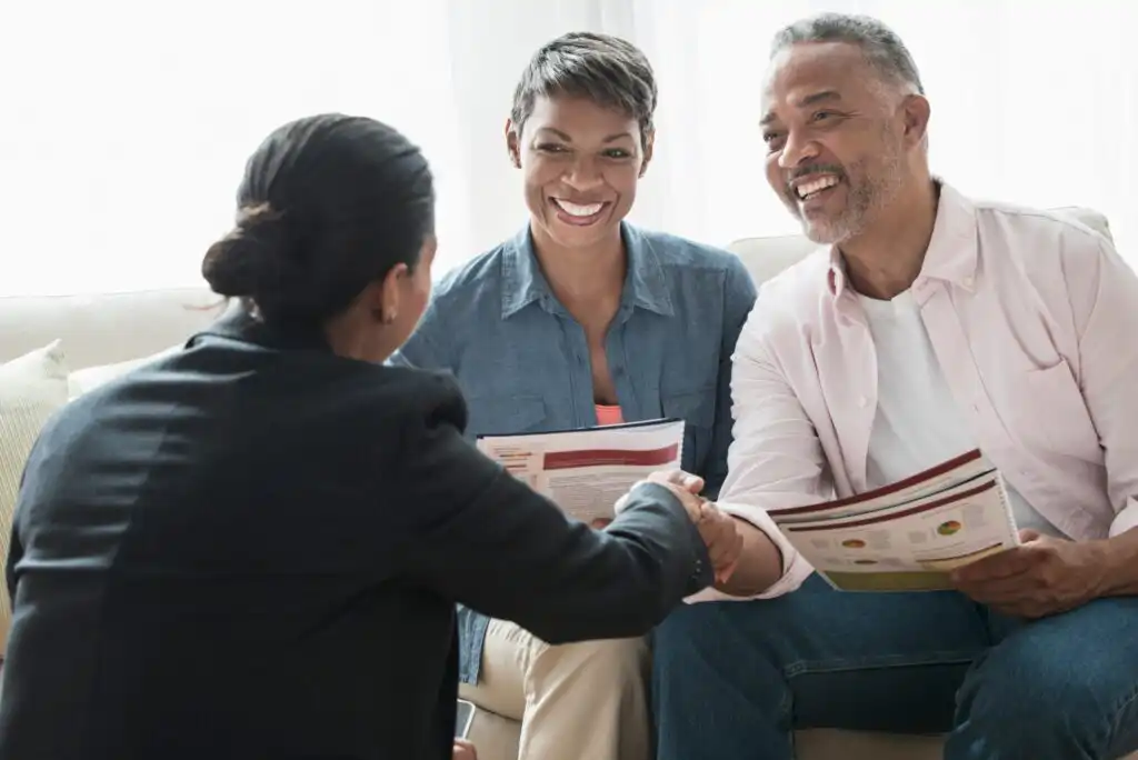 A professional woman shakes hands with a smiling man while another woman sits beside him, all holding documents and sitting on a couch, suggesting a positive business or financial meeting in a bright room.