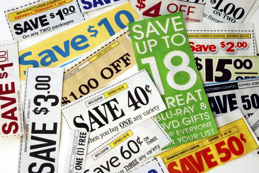 A variety of colorful paper coupons offering discounts such as “Save $10,” “Save $2.00,” and “Save 40¢” are spread out, overlapping each other. The coupons feature bold text and different designs.
