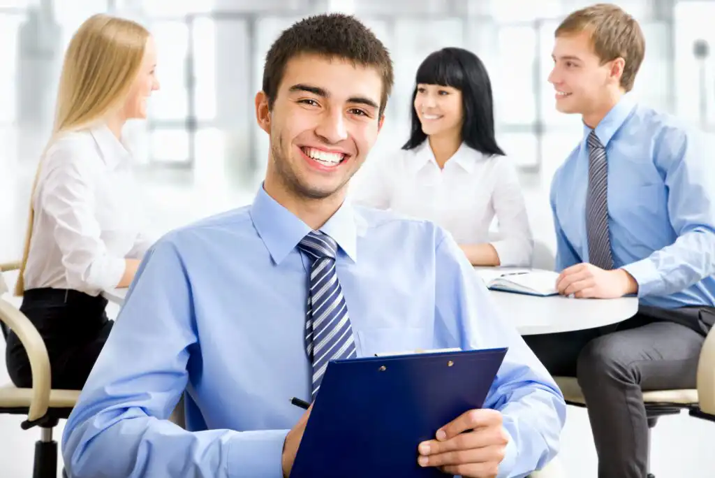 A young man in a blue shirt and striped tie smiles while holding a clipboard, with three colleagues having a discussion at a table in the background in a bright office setting.