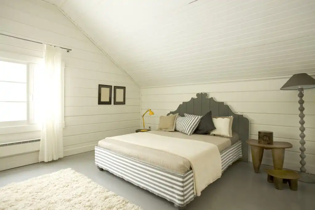 A cozy bedroom with white paneled walls and ceiling, a large bed with striped base, neutral bedding and pillows, two side tables, a tall gray lamp, framed pictures, white curtains, and a plush cream rug.