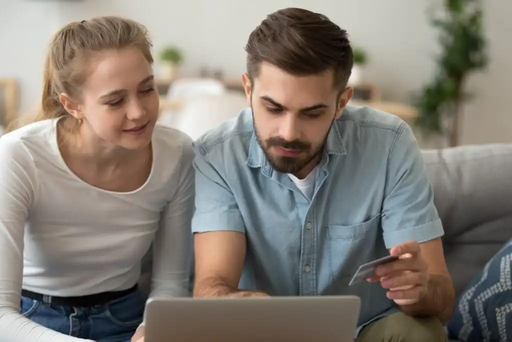 A young couple sits on a couch, looking at a laptop together. The man holds a credit card, and both appear focused and engaged with the screen, suggesting they are shopping or managing finances online.