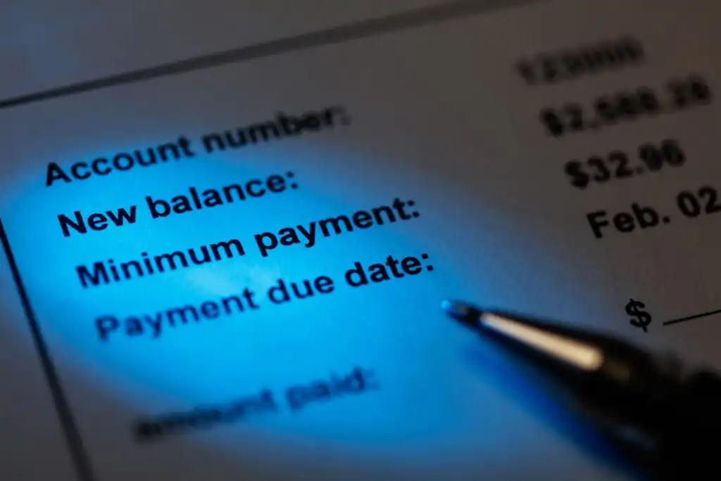 A close-up of a financial statement under blue light showing account details, including New balance, Minimum payment, and Payment due date, with a pen tip pointing at the paper.