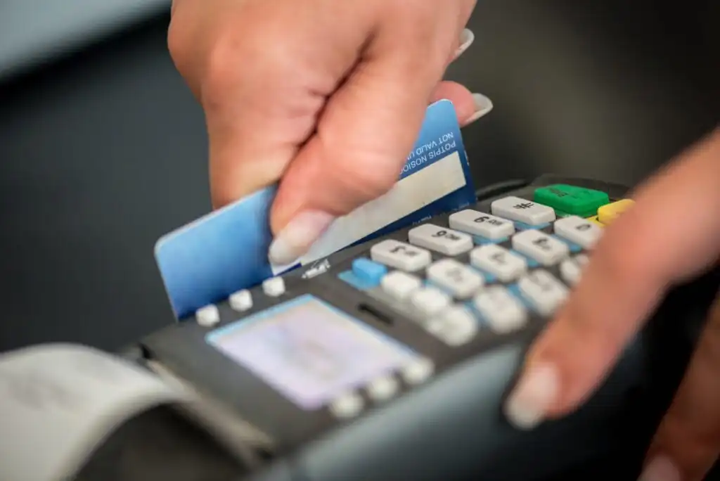 A close-up of a persons hand swiping a blue credit card through a payment terminal with a keypad and display screen.