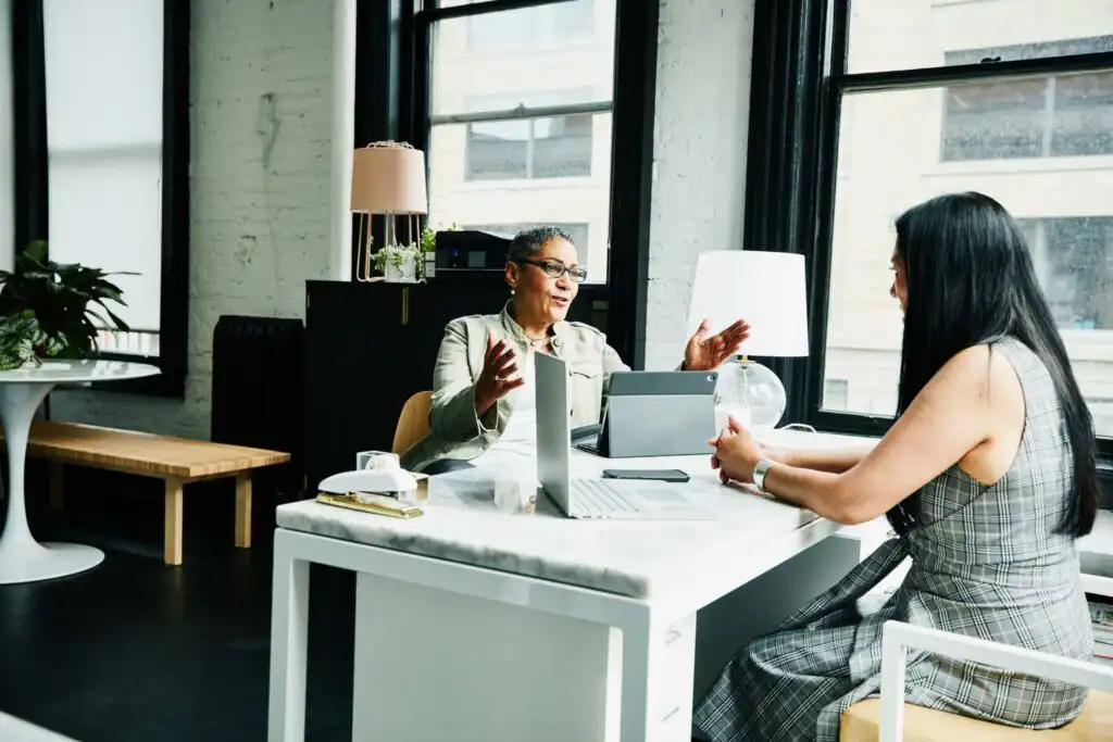 Two women sit across from each other at a modern office desk, engaged in conversation. One gestures with her hands while the other listens. Laptops, papers, and lamps are on the desk; large windows provide natural light.