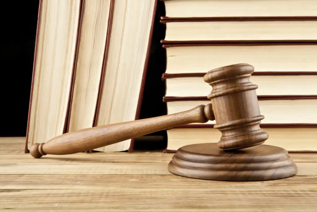A wooden judges gavel rests on its sound block in front of a stack of large, hardcover law books on a wooden surface.