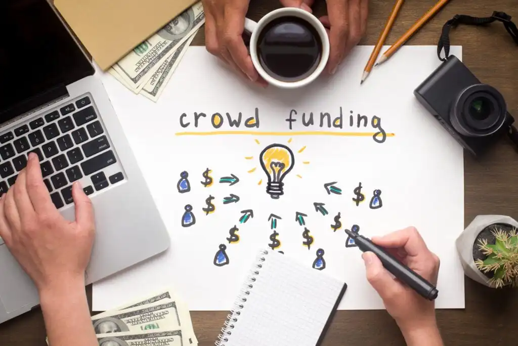 Top view of a desk with a laptop, cash, coffee, camera, notepad, and a hand drawing a “crowd funding” concept: a lightbulb in the center with arrows, people icons, and dollar signs around it.