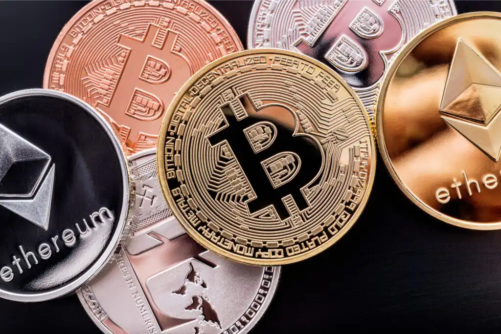 Close-up of physical coins representing cryptocurrencies, including gold and silver Bitcoin coins and Ethereum coins with their logos and names visible, arranged on a dark surface.