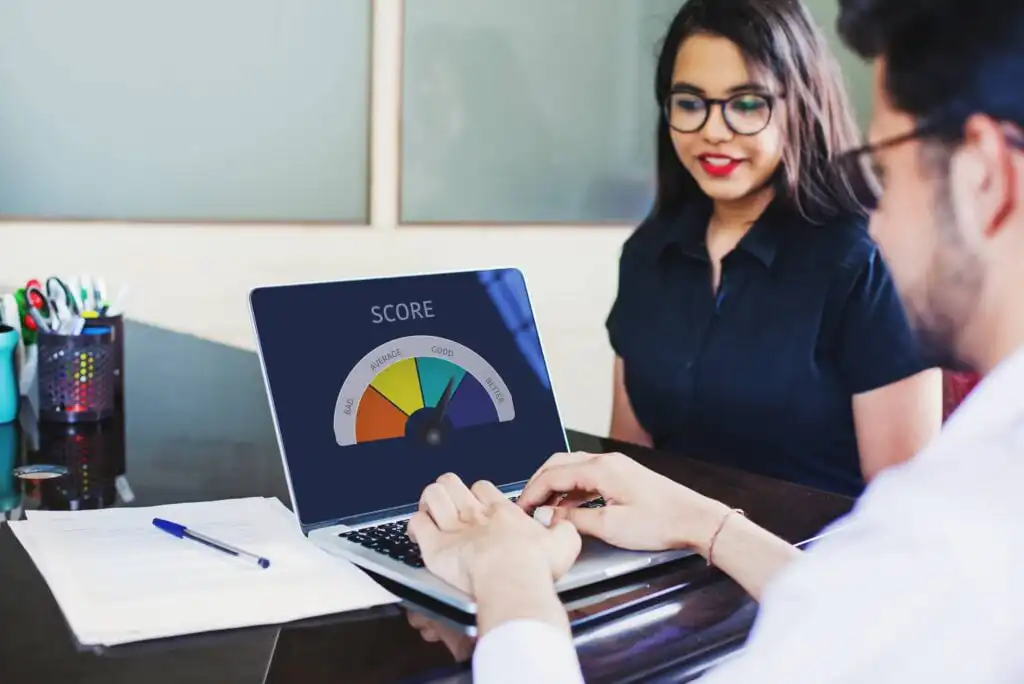 A person sits across from another at a desk, looking at a laptop displaying a credit score gauge in the Poor range. Papers and a pen are on the desk.