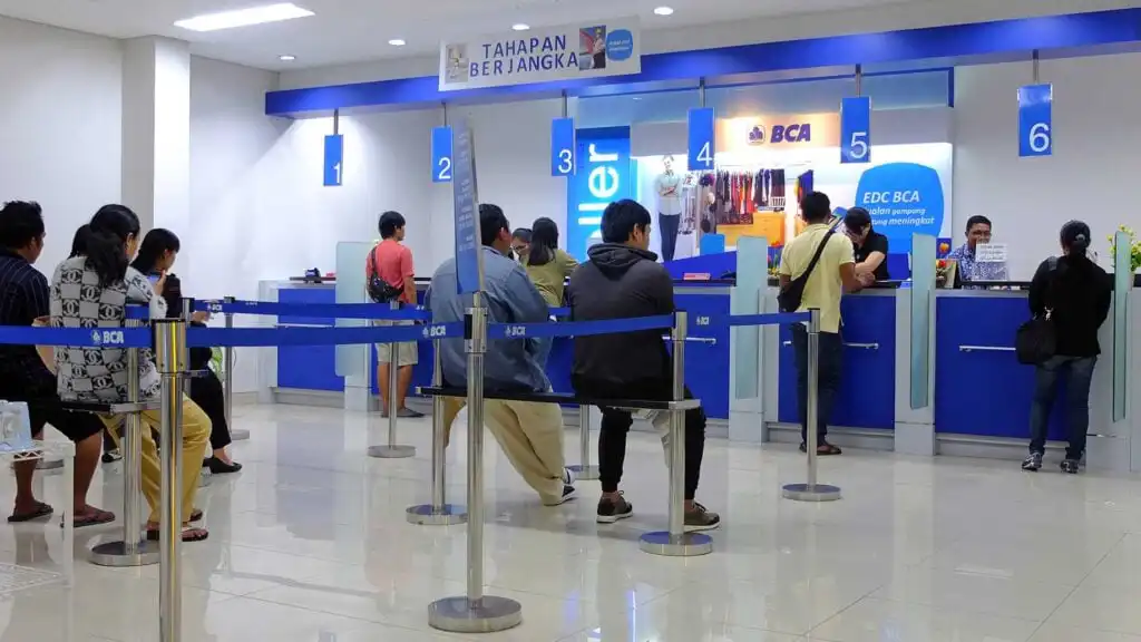 People are seated and standing in line at a bank counter labeled BCA, waiting for their turn at numbered service desks. Blue and white signs and partitions separate the queuing area.