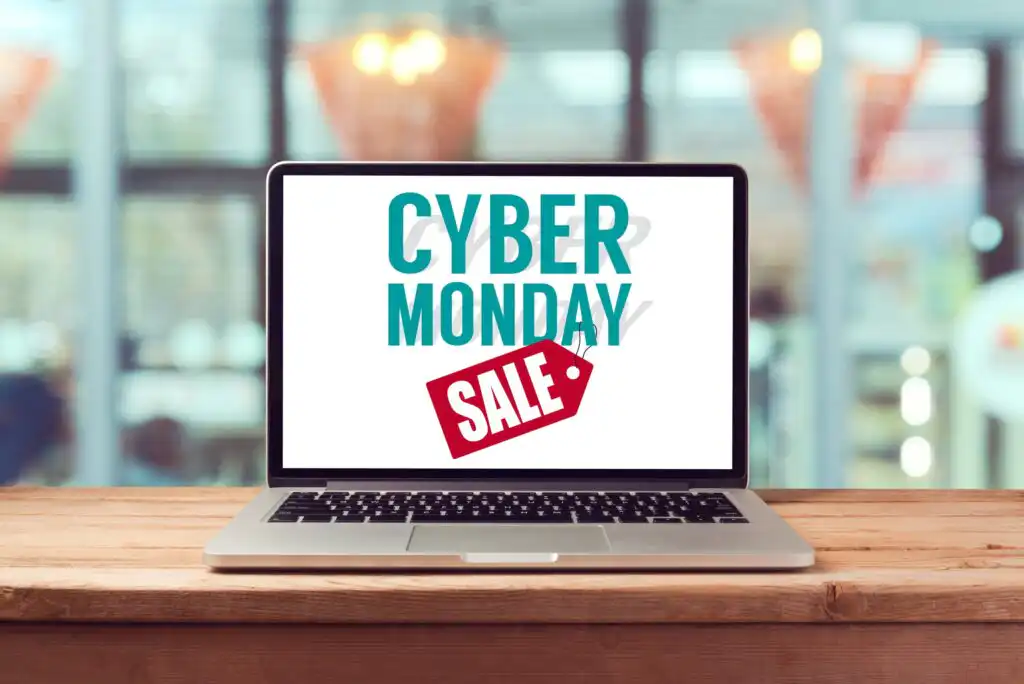 A laptop on a wooden table displays the words CYBER MONDAY SALE on its screen, with the word SALE inside a red tag. The background is blurred, showing large windows and hanging lights.