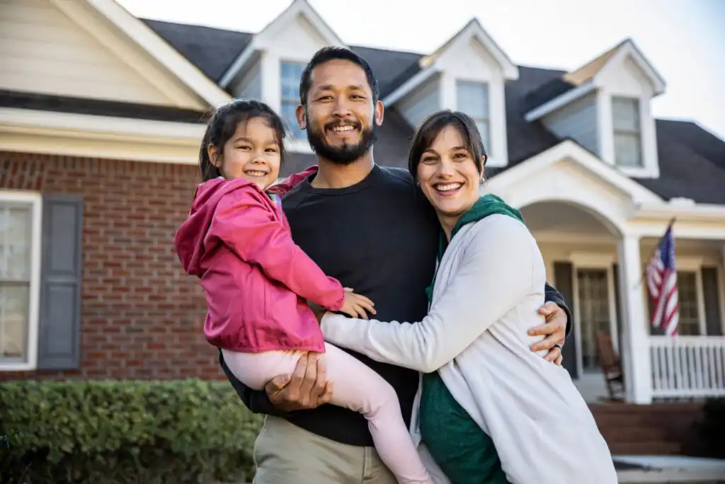 A smiling family of three—father, mother, and young daughter—stand together outside a brick house with white trim and an American flag, posing happily for the camera.