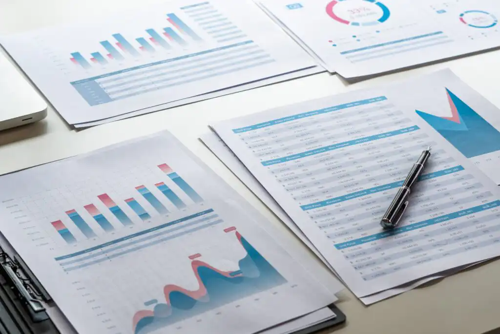 Several sheets of paper with colorful bar graphs, line charts, and tables are spread on a white desk. A pen rests on one sheet, indicating work involving business analytics or financial data analysis.