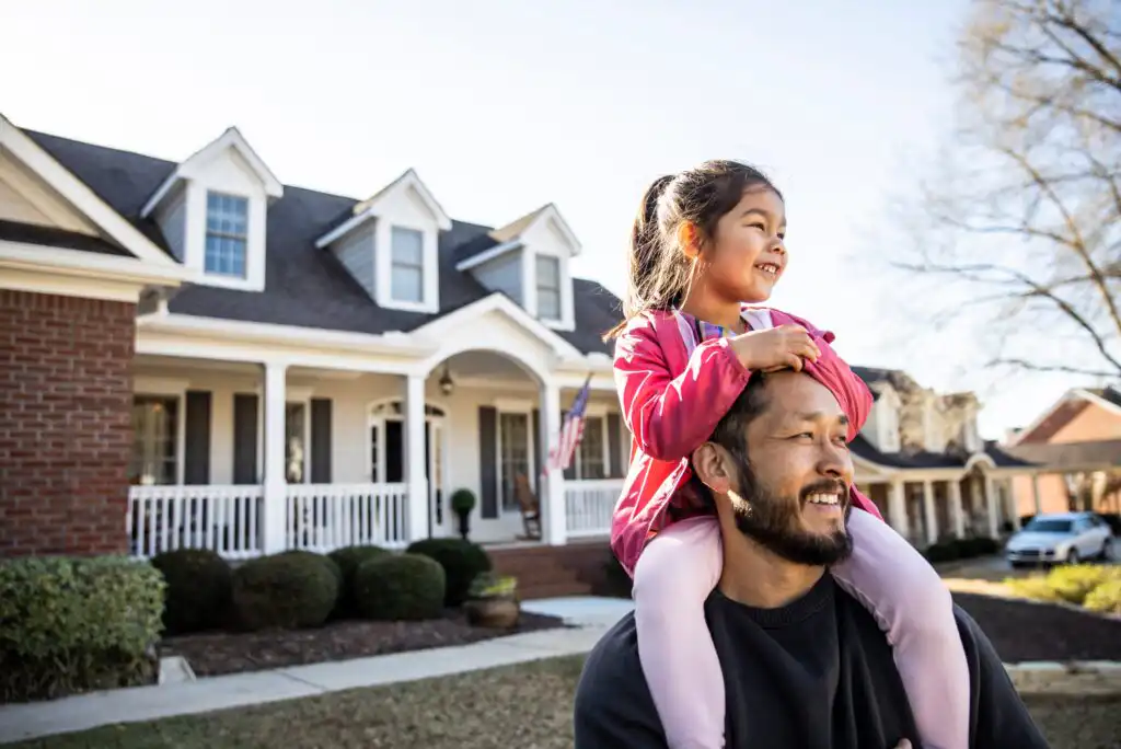 A smiling man carries a young girl on his shoulders in front of a suburban house with a porch and a well-kept yard on a sunny day. The girl is laughing and both appear happy.